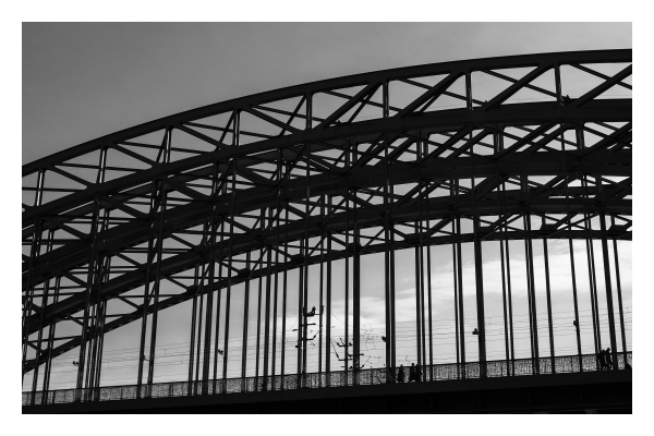 Schwarz-weiß Foto im Querformat. Ausschnitt einer Bogenbrücke. Diese besteht aus einem Metallgerüst. Es sind also ganz viele kurze und lange schwarze Linien, die sich gegen den Himmel abzeichnen. 