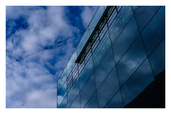 Foto im Querformat. Blick von unten an einem modernen Bürogebäude hoch. Dieses nimmt den rechten Teil des Bildes ein, das Gebäude hat eine verspiegelte Fassade, die aus vielen gleich großen Rechtecken besteht. Weiter oben sind rechteckige Fenster, die ebenfalls spiegeln. In allem spiegelt sich der bewölkte Himmel, welcher den linken Teil des Bildes einnimmt. Himmel und Gebäude sind beide in einem dunkelblauen Farbton getaucht. 