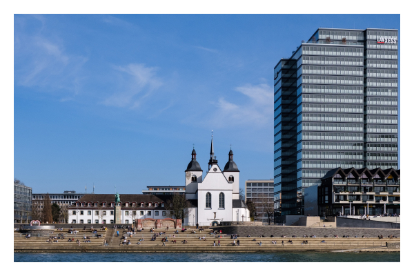 Foto im Querformat. Blick über den Rhein aufs andere Ufer. Rechts ist ein riesiges modernes Bürogebäude. Mittig ist eine, im Vergleich dazu, kleine weiße Kirche mit zwei Türmen. Davor sind große Steintreppen auf denen Menschen sitzen. Der Himmel ist leicht bewölkt. 