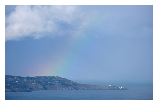 Foto im Querformat. Teleaufnahme seitlich auf eine Landzunge. Rechts ist das Meer, links ragt die Küste ins Bild. Über der Landzunge ist ein Regenbogen. Der Himmel ist bewölkt, über der Landzunge ist eine große weiße Wolke. 