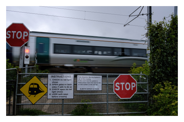 Foto im Querformat. Ein Bahnübergang. Gerade fährt ein Zug durch. Vor dem Bahnübergang ist eine Absperrung mit zwei roten stop Schildern. Zusätzlich sind noch Schilder darauf mit folgendem englischen Text:
STOP
INSTRUCTIONS
1. CHECK no red light shows
2. OPEN gates on both sides if safe to do so
3. CHECK again no red light shows
4. LOOK both ways before crossing
Train Detection Warning System in
use at this crossing.
In the event that the system is
faulty please report the fault to the
Lo Call number on the red sign.
ALWAYS EXPECT A TRAIN
STOP