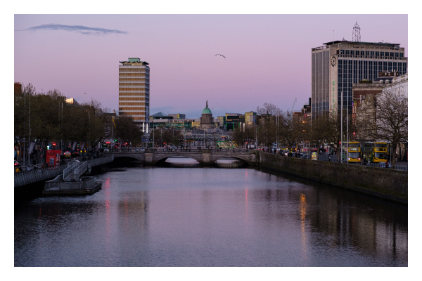 Foto im Querformat. Blick von einer Brücke entlang des Flusses Liffey in Dublin. Es ist Abend, der Himmel ist rötlich blau. Links und rechts sind hohe Gebäude, die sich teils im Wasser spiegeln. Direkt geradeaus ist ein kleineres altes Gebäude mit grünem runden Dach. Mittig im Bild über dem Fluss fliegt eine Möwe. 