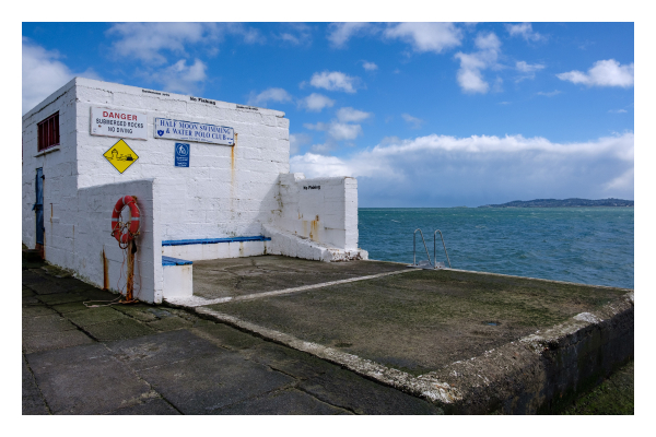 Foto im Querformat. Ein asphaltiertes Pier am Meer. Links ist eine weiße Gebäude aus Stein, daran sind einige Schilder und ein Rettungsring. Zum Meer hin ist das Geländer einer Leiter im Bild. Das Meer ist blau, am Horizont ist eine grüne Landzunge. Der Himmel ist leicht bewölkt. 
Auf den Schildern steht auf Englisch:
No Fishing
DANGER
SUBMERGED ROCKS
NO DIVING
HALF MOON SWIMMING & WATER POLO CLUB 
