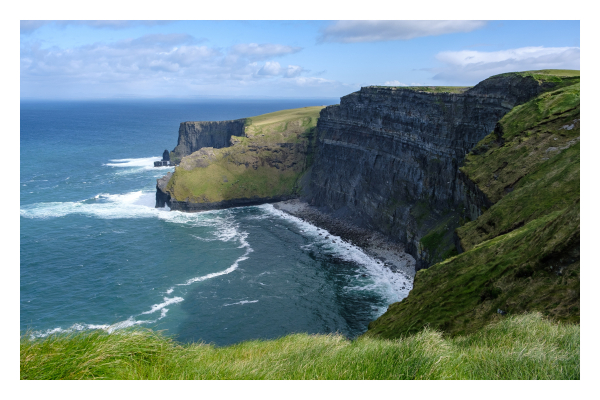 Foto im Querformat. Blick auf die Cliffs of Moher, eine Steilküste in Irland. Eine riesige Felswand ragt von rechts ins Bild. Sie beschreibt einen Bogen, unten ist ein kleiner Kiesstrand. Am Ende des Bogens ragt wieder ein großer Felsen weit ins Meer. Das Meer unten ist weiß an den Felsen, sonst blau. Ganz im Vordergrund ist Gras, leicht unscharf. Der Himmel ist leicht bewölkt. 