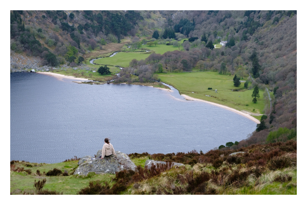 Foto im Querformat. Blick auf einen See von einer Anhöhe. Die Anhöhe ist mit grünem Gras und braunen Pflanzen bewachsen. Auf einem grauen Felsen sitzt eine Person und schaut herab zum See. Der See hat einen weißen Strand, dahinter sind grüne Wiesen, ein Fluss fließt in den See und ein Anwesen ist hinter Bäumen zu sehen. 