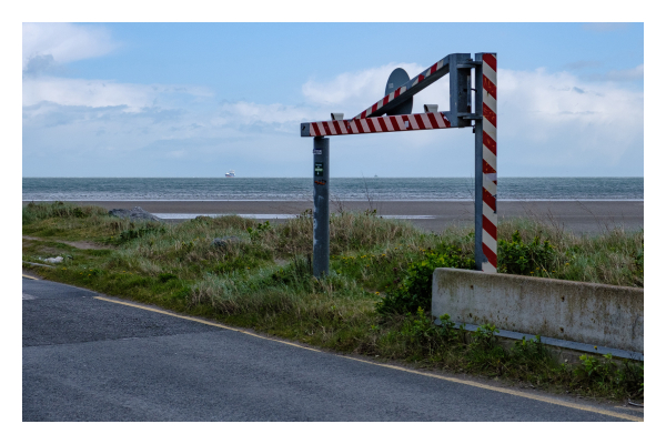 Foto im Querformat. Ein Teil einer Straße, daneben ist Gras, dahinter Strand und schließlich das Meer. Im Gras ist eine Höhenbeschränkungsschranke, die momentan geöffnet ist. Durch die zwei Pfosten sieht man ein Schiff am Horizont. Links neben der Schranke ist ebenfalls ein Schiff am Horizont. Die Schranke ist rot weiß gestreift, der Himmel ist leicht bewölkt. 