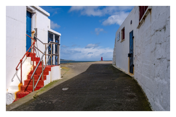 Foto im Querformat. Blick entlang eines sehr langen Stegs, vorne sind links und rechts jeweils ein weißes Gebäude aus Stein. In beiden ist eine blaue Türe, am linken führt zu dieser eine rote Treppe um die Ecke. Ganz am Ende des Stegs, am Horizont, ist ein roter Leuchtturm. Auf die Entfernung sieht dieser sehr winzig aus. Links ist ein Teil vom Meer, der Himmel ist blau und leicht bewölkt. 