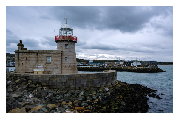 Foto im Querformat. Der Leuchtturm im Hafen von Howth in Irland. Ein Turm aus hellbraunem Stein mit einem Gebäude daran. Der Turm hat oben ein rotes Geländer und eine weiße Kuppel mit Außendruck Fenstern. Der Blick ist ins Landesinnere, hinter dem Turm ist ein Hafenbecken und ein Pier. Der Himmel ist bedeckt. 