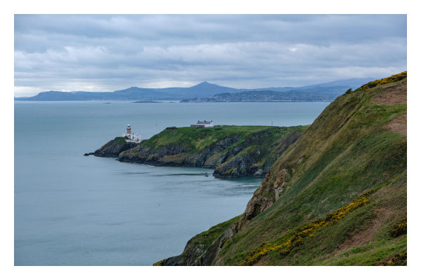 Foto im Querformat. Blick einer Landzunge entlang, die ins Meer ragt und immer niedriger wird. Ganz am Ende ist ein weißer Leuchtturm. Die Landzunge beschreibt einen Bogen nach links. Hinter der Landzunge ist Meer und am Horizont ist Festland. Der Himmel ist bedeckt. 