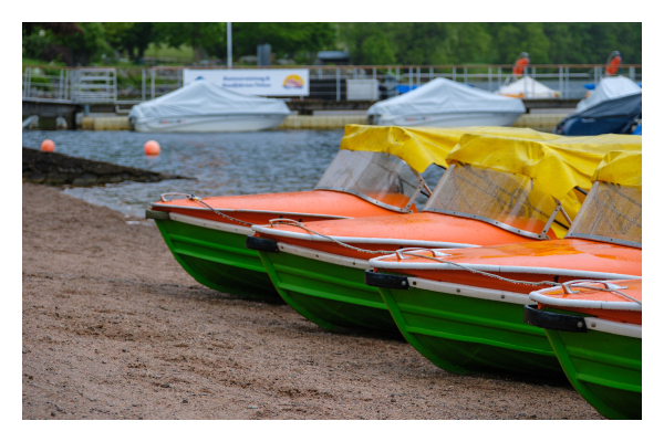 Foto im Querformat. Ein Strand, mehrere kleine Boote liegen am Ufer nebeneinander und sind durch ein Kabel aneinander gebunden. Alle Boote sehen gleich aus, grüner Rumpf, oben orange, eine Windschutzscheibe aus Glas und eine gelbe Plastikfolie als Dach. Die Boote ragen von rechts ins Bild. 