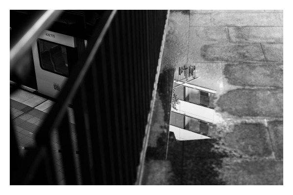 Schwarz-weiß Foto im Querformat. Blick Richtung Boden an einem Bahnhof. Mittig geht ein Geländer von oben nach unten durchs Bild. Rechts davon ist am Boden eine Pfütze, in der sich die Fenster eines Hauses spiegeln. Links vom Geländer geht der Blick ein Stockwerk tiefer aufs Gleis. Ein kleiner Ausschnitt der S-Bahn ist zu sehen. Auf den ersten Blick sieht es so aus, als ob sich die Bahn in der Pfütze spiegeln würde. 