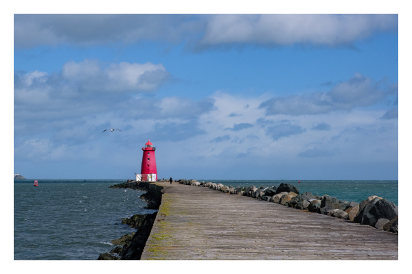 Foto im Querformat. Blick entlang eines breiten Steges aus Stein. Links und rechts ist Wasser. Ganz am Ende ist ein knallroter Leuchtturm, der unten weiß ist. Eine einzelne dunkel gekleidete Person läuft Richtung Leuchtturm. Links fliegt eine Möwe. Der Himmel ist leicht bewölkt. 