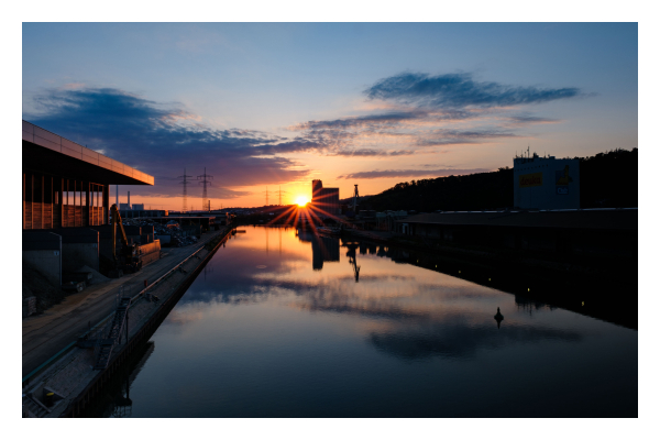 Foto im Querformat. Sonnenuntergang. Blick entlang eines Flusses, an den Ufern sind Industrieanlagen. Hinten rechts erhebt sich ein Gebäude, dahinter geht die Sonne unter. Die Sonne erscheint wie ein Stern mit strahlen. Der Himmel ist orange bis blau und leicht bewölkt. Er spiegelt sich im ruhigen Fluss. Die Sonne taucht das linke Ufer in rötliches Loch, das rechte Ufer versinkt im Schatten. 