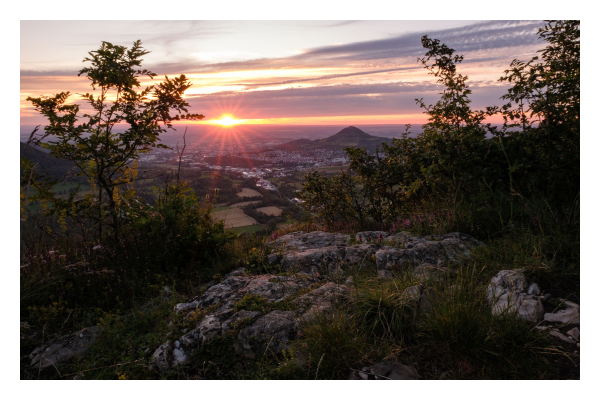 Foto im Querformat. Sonnenuntergang. Blick von einem hochgelegenen Felsen über das Tal. Im Vordergrund ist das Felsenplateau, links und rechts sind Pflanzen, die den Blick ins Tal einrahmen. Im Tal sind Felder, Wiesen und eine große Stadt. Rechts ist ein Berg, der dominant hervorsticht. Am Horizont ist eine dicke Wolke, die Sonne ist genau zwischen dieser Wolke und dem Horizont. Sie strahlt wie ein Stern. Der restliche Himmel ist teils klar, teils bewölkt. 