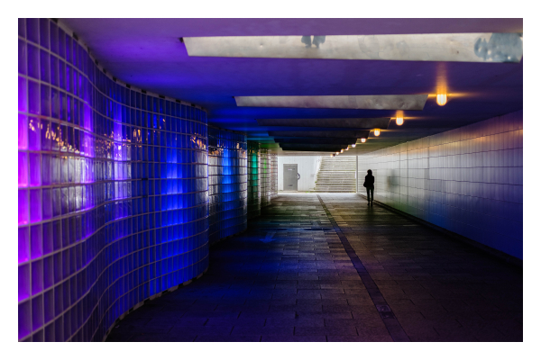 Foto im Querformat. Blick in eine Unterführung. Die linkes Wand besteht aus Glasfliesen, die von hinten beleuchtet sind. Vorne violett, nach hinten dann blau und schließlich grün. Außerdem ist die Wand wellenförmig. Die andere Seite der Unterführung ist eine gerade, helle Wand. An der Decke sind in regelmäßigen Abständen gelbe Lichter, die zum Ausgang der Unterführung gehen. Dieser ist sehr hell im Vergleich zur dunklen Unterführung. Am Ende der Unterführung läuft ganz rechts eine einzelne Person, diese ist nur eine Silhouette. Sie wirft einen Schatten an die rechte Wand. 