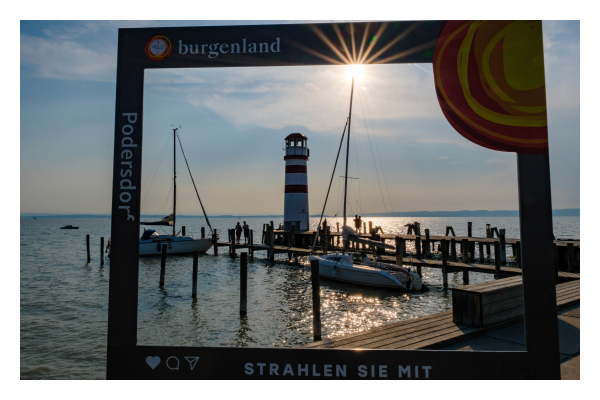 Foto im Querformat. Blick auf einen Steg an einem See. Kleine Segelboote liegen an, am Ende des Stegs ist ein kleiner rot weiß gestreifter Leuchtturm. Auf dem Steg sind einige Menschen, die aber nur als Silhouette zu erkennen sind. Im Vordergrund ist eine Art Bilderrahmen, durch die die ganze Szene zu sehen ist. Die Sonne ist genau am oberen Teil des Rahmens und bildet einen Sonnenstern. Auf dem Rahmen steht Burgenland, Podersdorf und „strahlen sie mit“. Der Himmel ist leicht bewölkt. 