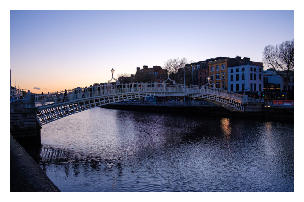 Foto im Querformat. Blick auf die Ha'penny Bridge in Dublin, Irland. Die Brücke ist eine Fußgängerbrücke aus weiß lackiertem Gusseisen. Sie ist leicht gebogen und geht über den Fluss Liffey. Es ist Abend, links am Horizont ist die Sonne gerade untergegangen und der Himmel ist rötlich, weiter oben blau und klar. 