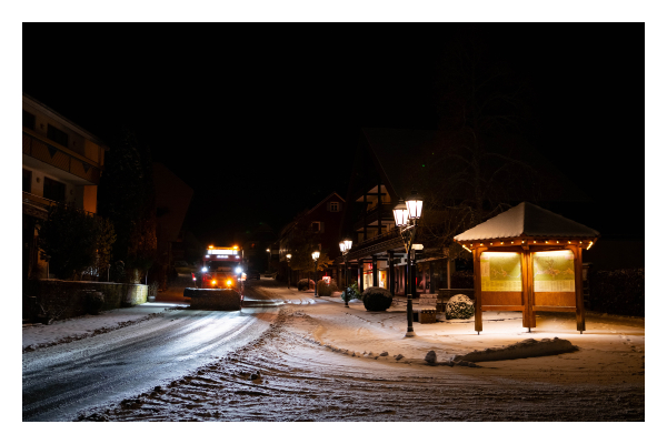 Foto im Querformat. Blick über eine verschneite Straßenkreuzung in einer Stadt. Rechts sind Straßenlaternen und eine überdachte informationstafel. Diese ist beleuchtet. Auf der Straße kommt gerade ein Schneepflug angefahren, dieser ist ebenfalls hell beleuchtet. Der Himmel darüber ist komplett schwarz. 