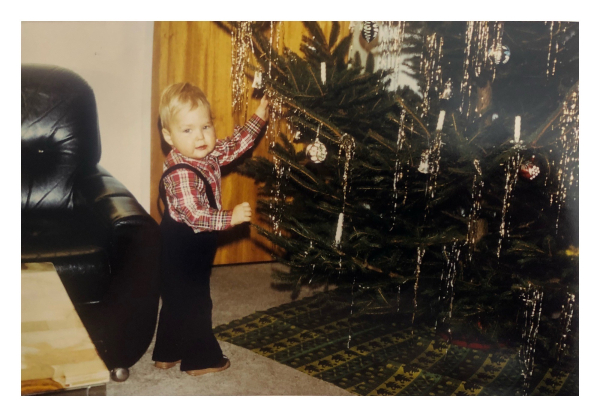 Foto im Querformat. Ich, Ende der 70er Jahre, vor einem geschmückten Weihnachtsbaum. Am Baum hängen Kugeln, echte Kerzen und Lametta. Ich trage eine dunkelblaue Latzhose mit Schlag und ein rot weiß kariertes Hemd. Meine Pausbacken sind aus einer anderen Welt und auf meinem Kopf prangt güldenes Haar. Ich schaue direkt in die Kamera, während ich am Baum mit der linken Hand rumfummle. Meine Mutter schimpft vermutlich von hinter der Kamera. 