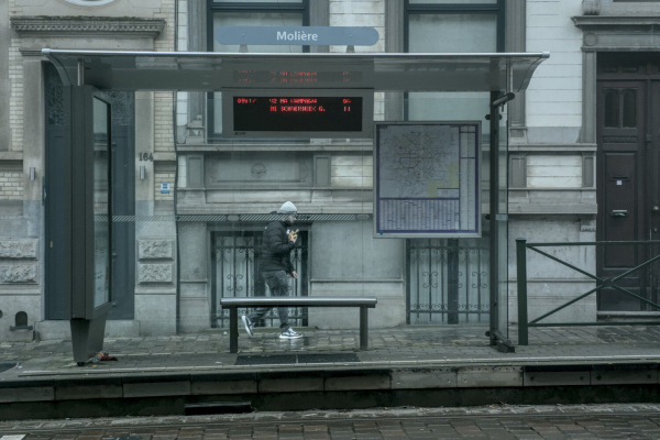 Person walking past a tram stop, he is visible through the glass pane of the stop. His eyes are covered by a line on the glass pane.