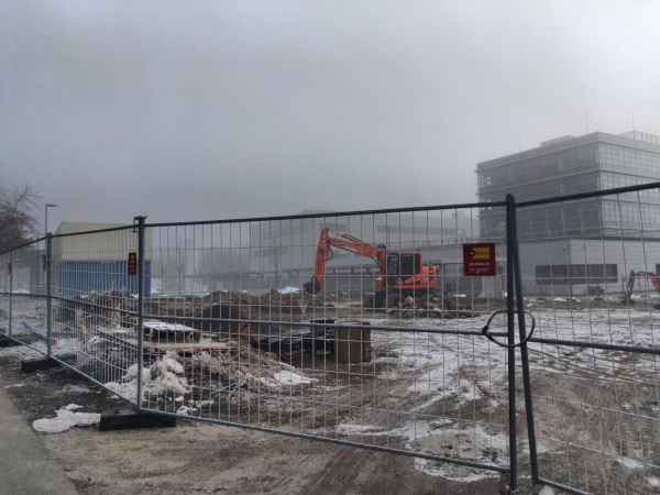 Ein mehrstöckiges Gebäude vor sehr grauem Himmel, im Vordergrund eine Schotterfläche mit Fahrzeugspuren, die durch einen Bauzaun abgesperrt ist. Auf der Schotterfläche stehen ein Bagger und mehrere Container. 