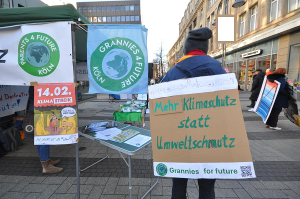 Das Bild zeigt den Aufbau der Kundgebung "Gehe wählen! Wähle demokratisch! Wähle Klimaschutz" in Köln, veranstaltet von verschiedenen Kölner Klimaschutzgruppen. Im Vordergrund steht eine Person mit einer selbstgemachten Papptafel auf dem Rücken, auf der „Mehr Klimaschutz statt Umweltschmutz“ steht. Die Person trägt eine dunkle Jacke und eine Mütze.  Im Hintergrund sind weitere Aktivist*innen zu sehen, Infostände mit Plakaten von „Parents for Future Köln“ und „Grannies for Future Köln“ und ein Plakat der Firdays for Future Köln mit dem Datum „14.02. Klimastreik“. Die Stände befinden sich auf der Breiten Straße in Köln. Bild: Ulla Kellerwessel, nicht für kommerzielle Zwecke