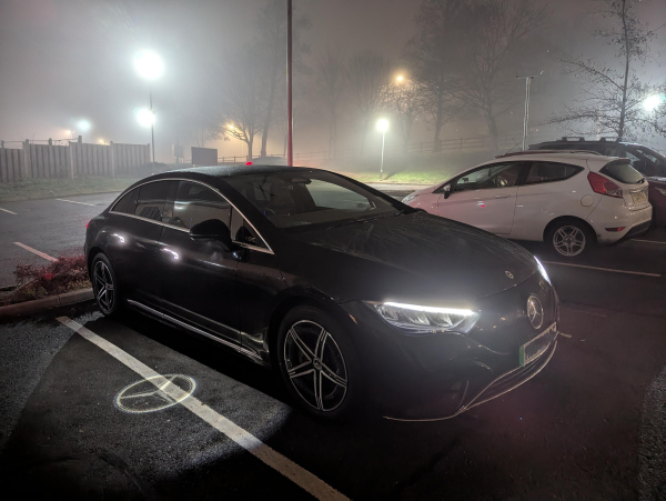 Photo of a Mercedes EQE electric sedan parked at night in the fog. The puddle lights under the wing mirrors are projecting the Mercedes logo onto the ground next to the car.