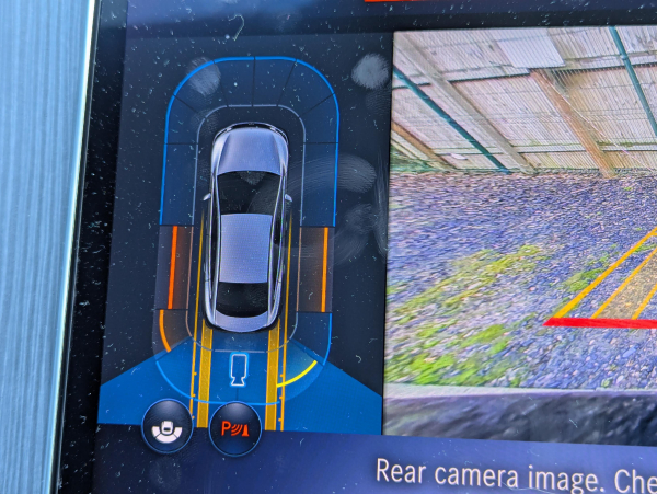 Closeup photo of the central infotainment screen inside a Mercedes EQE showing the parking sensor status display, with orange proximity warnings on both sides of the car.