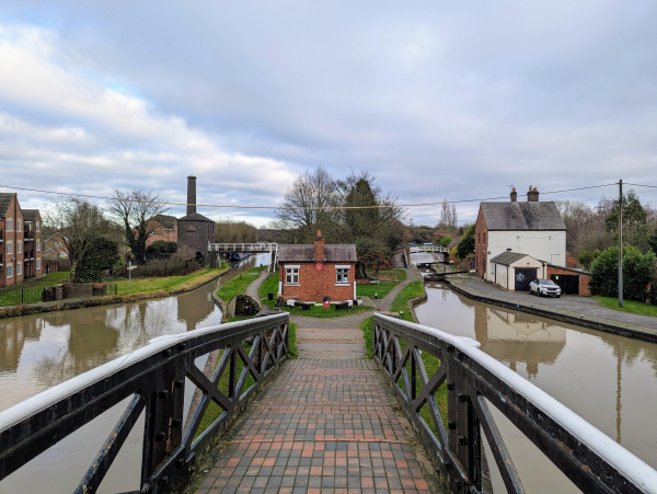 Photo of the view along a foot bridge towards a peninsula of land between two canals. Alongside the canal on the left of an old bright engine house with a prominent chimney, and on the canal on the right is a lock and beside it a lock keeper's cottage.