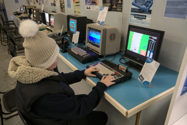 Photo of a long desk with a dozen or so old 8-bit computers set up on it, with a child sitting at the nearest one playing a primitive car racing game.