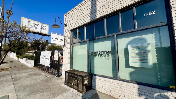 Image of a Stumptown Coffee Roasters location featuring a large sign, a billboard reading "Welcome to HomePlate," and a poster in the window. The building has a modern brick facade, and the street is lined with trees and sidewalks.