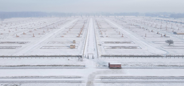 A wide aerial view of a snow-covered landscape at the former Auschwitz II-Birkenau camp. Rows of rectangular building markings and brick chimneys extend symmetrically, marking the remains of structures. Railway tracks run in the foreground, with a lone red freight car positioned nearby. A single tree and barbed wire fences frame the stark and desolate scene, evoking a somber and reflective mood.