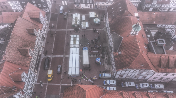 An aerial view of a town square featuring several market stalls and a gathering of people. The scene includes various vehicles, including police cars, and is surrounded by traditional buildings with red-tiled roofs. A misty atmosphere gives the image a subdued tone.