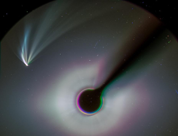 A picture of a comet with several tails seen in the field of view of a spacecraft camera. There is a black stick with a round circle at the end of it which blocks the Sun, allowing faint nearby things to be seen. The image has greens and purples in it due to the shifts and rotations introduced to make the moving comet line up in the three sequential black and white images added to make the colour combination.