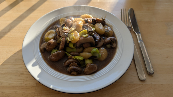 Auf einem weißen Teller sind vegane Gnocchi mit Pilzen und Lauch, in dunkler Soße.

Daneben ein Besteck und alles zusammen auf einem Holztisch, der schräg von vorne von der Sonne beschienen wird.