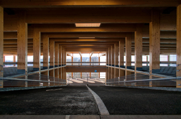 View down the blacktop covered central ramp of an empty parking garage connection two decks. It is build from massive wooden glulam beams and pillars. At the end of the ramp the outer wall opens up and sun creates shadows on the floor.