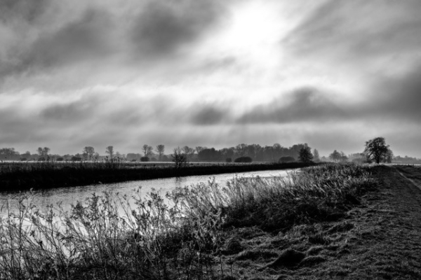 Eine schwarz-weiße Landschaft mit einem gewundenen Fluss, der von Vegetation gesenkt ist, unter einem dramatischen Himmel mit Lichtstrahlen, die durch die Wolken brechen. Silhouettenbäume und Felder sind im Hintergrund sichtbar.