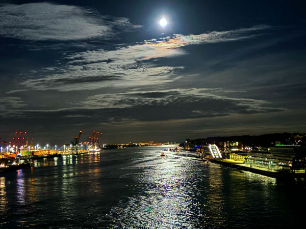 Der Hamburger Hafen mit seinen Verladeanlagen links und rechts der Elbe. Dunkler Nachthimmel, mit ein paar Wolken, welche von Vollmond illuminiert werden. Der Mond und die Lichter der Hafenanlagen spiegeln sich auf dem dunklen Wasser.