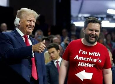 Horizontal color photo.
Trump with maxi pad on his ear (left) and running mate JD Vance (right) at a campaign event. Both are grinning. Trump is giving a thumbs up, Vance is wearing a red T-shirt with the words, "I'm with America's Hitler," and an arrow pointing to Trump.