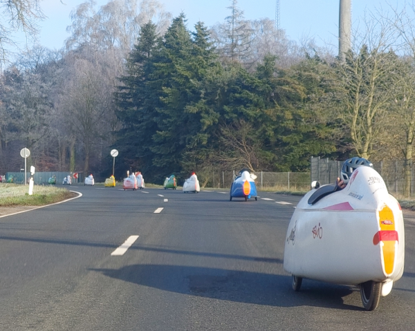 Eine Gruppe Velomobile in verschiedenen Farben rollt eine lange Linkskurve in der Sonne entlang. Im Hintergrund Nadelbäume und reif besetzte Laubbäume. 