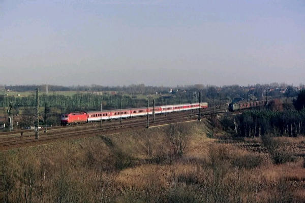 Das Bild zeigt eine weitläufige Landschaft mit mehreren parallel verlaufenden Eisenbahngleisen. Auf dem linken Gleis fährt ein roter Intercity-Zug mit mehreren weißen Personenwagen, der von einer modernen Elektrolokomotive gezogen wird. Rechts daneben ist ein langer Güterzug mit einer grünen Lokomotive zu sehen. Im Hintergrund erstrecken sich offene Felder und eine lockere Ansammlung von Häusern. Der Himmel ist klar und blau, was auf einen sonnigen, kalten Tag hindeutet. Im Vordergrund sind kahle Büsche und Grasflächen sichtbar, die den Eindruck eines ländlichen Gebiets verstärken.

