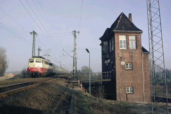 Das Bild zeigt eine alte Elektrolokomotive der Baureihe 103 in creme-roter Lackierung, die auf einem leicht erhöhten Bahndamm mit einem Zug vorbeifährt. Links und rechts des Gleises sind Oberleitungsmasten mit elektrischen Leitungen zu sehen. Rechts im Vordergrund steht ein markantes, backsteingemauertes Stellwerk mit einem spitzen Ziegeldach und mehreren Fenstern. Eine schmale Metallbrücke führt vom Stellwerk über einen Graben zu den Gleisen. Die Umgebung wirkt ländlich, mit vereinzelten Bäumen im Hintergrund und einem bewölkten, aber hellen Himmel. Die Atmosphäre deutet auf einen klaren, kalten Tag hin.

