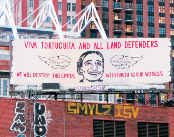 Mural covering a billboard in a cityscape. It’s an illustration of Tortuguita’s smiling face between two wings with his birth and death dates written on them. The sign reads “viva Tortuguita and all land defenders. We will destroy this empire with earth as our witness.”