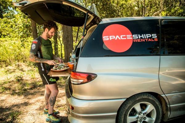 Space Traveller cooking campsite tacos in Australia. Campervan is shown half and the Space Traveller is standing at the back where the cooking area is located and is using the included cooking gear to create a simple yet tasty dish. Trees and forrest in the background.