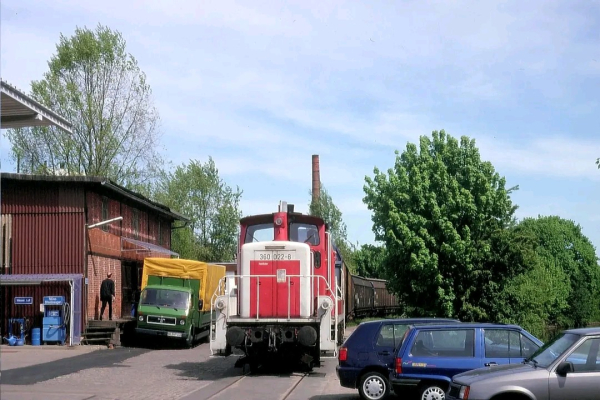 Das Bild zeigt eine rote Rangierlokomotive mit der Nummer "360 022-8", die auf einem Gleis in einem Industrie- oder Lagerbereich steht. Links im Bild befinden sich ein alter Ziegelschuppen und ein grüner Lieferwagen mit gelber Plane. Im Vordergrund sind parkende Autos zu sehen, darunter ein blauer und ein grauer PKW. Im Hintergrund sind grüne Bäume und ein hoher, schmaler Schornstein erkennbar. Der Himmel ist klar und blau.

