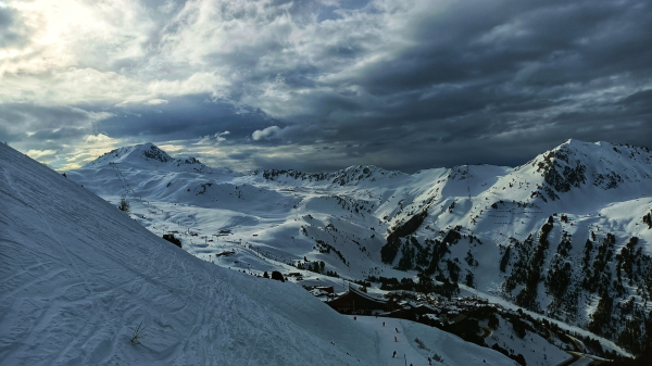 photo d'un paysage de montagne enneigé. Elle monte un vallon autour duquel on distingue des pistes et des remontées mécaniques. Le ciel est chargé de nuages qui assombrissent l'atmosphère, même si le soleil arrive à percer sur la gauche de l'image. l'ensemble donne une touche dramatique à l'image.