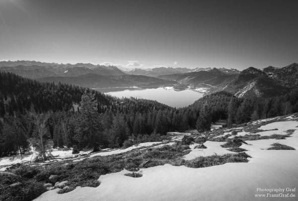 A stunning black and white photograph captured by Photography Graf showcases a serene winter landscape. The image features a snowy mountain towering in the background, surrounded by tall trees and a tranquil lake in the foreground. The scene exudes a sense of tranquility and isolation, with the dominant colors of grey, black, and white creating a harmonious and peaceful atmosphere. The snow-covered mountain range in the distance adds to the rugged beauty of the wilderness. The clear sky with clouds overhead enhances the overall sense of calm and serenity in this monochromatic winter wonderland.