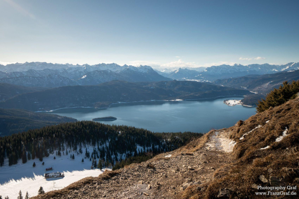 A stunning landscape featuring a majestic mountain standing tall against a clear blue sky. The mountain is covered in snow, creating a picturesque scene. At the base of the mountain, there is a serene body of water, reflecting the surrounding beauty. The snow-capped peak contrasts beautifully with the brown rocky terrain below. Sparse trees dot the landscape, adding a touch of greenery to the otherwise rugged terrain. The image exudes a sense of tranquility and wilderness, capturing the essence of nature's raw beauty. This scene is a perfect example of the harmony between mountains, water, and snow in the great outdoors.