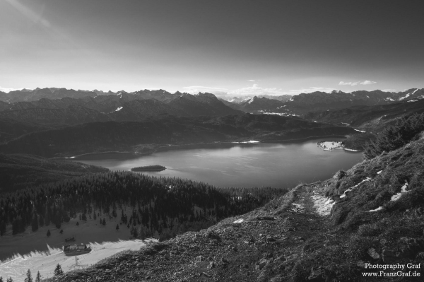 A stunning black and white landscape photograph depicting a serene lake surrounded by towering mountains. The dominant colors in the image are grey and black, creating a peaceful and tranquil atmosphere. The mountains in the background are snow-capped, adding to the majestic beauty of the scene. The sky is cloudy, adding a touch of drama to the composition. This highland wilderness setting exudes a sense of calm and solitude. The image is monochrome, emphasizing the natural beauty of the mountainous terrain and the reflective surface of the lake. The overall composition is breathtaking and evokes a sense of awe and wonder at the power and majesty of nature.