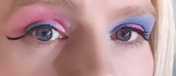 A closeup of Alice's bright blue eyes. They're wearing shimmering baby pink eyeshadow over their left eye and powder blue eyeshadow over their left eye. Each eye is lined on top with the opposite color of liquid eyeliner, and both have a bold black upper line and wing. Alice's blonde hair is framing the right side of the image.